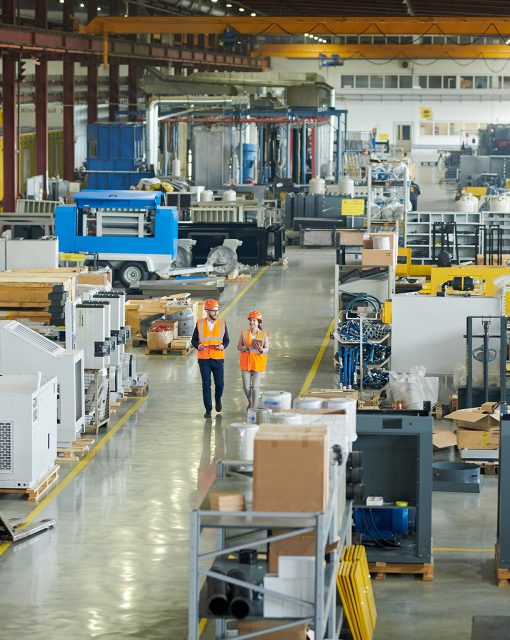 High angle full length portrait of bearded businessman wearing hardhat walking across production workshop accompanied by female factory employee, copy space